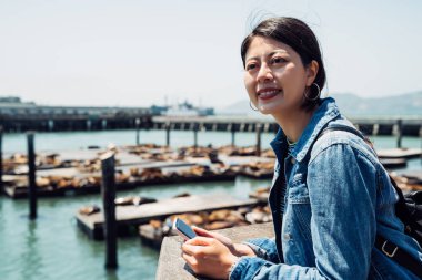 Asyalı kız holding cep telefonu pier 39 san Francisco'da gezi gülümseyerek. sevimli vahşi doğa hayvan deniz aslanları dinlenme fisherman wharf yüzen tahta gemide yalan üzerine güneş ışığı tadını çıkarın..