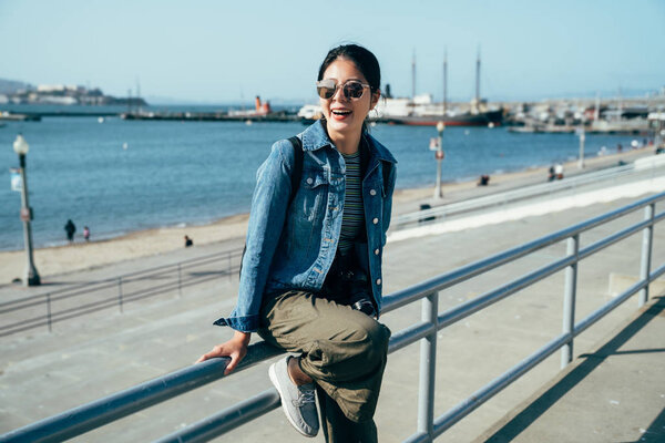 happy girl photographer wearing sunglasses enjoy the beautiful view of blue ocean and sky relying sitting on handrail near the port. young asian woman smiling laughing sightseeing in the pier 39.