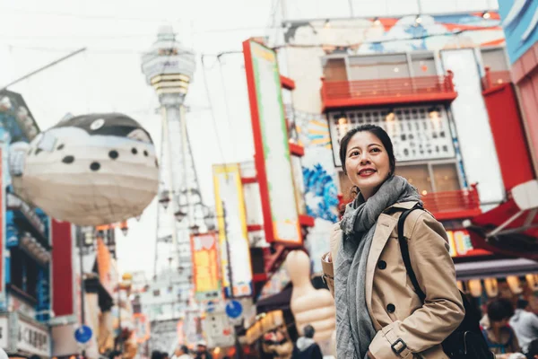 young female traveler cheerfully sightseeing standing on the teeming street in tsutenkaku on sunny day wearing scarf. huge puffer fish balloon flying in the sky. girl smiling looking around on road.