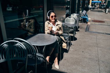 Güzel Asyalı kadın güneş gözlüğü açık havada sokak kafe kafede masada oturan kulaklıklar cep telefonunuzun müzik dinle. Genç kız rahatlatıcı restoranda güneş ışığı altında ücretsiz zamanında.
