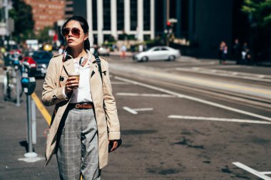 Asyalı kadın yürüyen açık havada sokak kahve fincanı güneş ışığı altında tutun. san Francisco'da sabah çalışacak genç hanım. güneş gözlüğü ve smart casual elbise giymiş şık iş kadını.