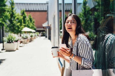 tarafından moda mağaza Vitrini mankenler ile duran genç tüketici kadın. Kağıt Poşetler tutarak ve açık havada neşeyle gülümseyen güvenerek güneşli bir gün boyunca smartphone kullanarak Asyalı kız.