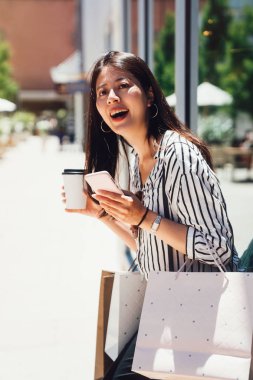 Şaşırtıcı gülümseyen güzel Asyalı kadın ayakta açık havada cep telefonuyla görüntü. Vitrin çanta ve neşeyle gülerek kahve tutan Giyim Mağazası dışında güvenerek dinlenme genç kız.