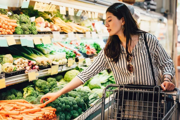 japanese woman in supermarket pushing shopping cart walking by the vegetables and fruits area. pretty asian lady picking up carrots beside broccoli purchase food health care. wife prepare for dinner