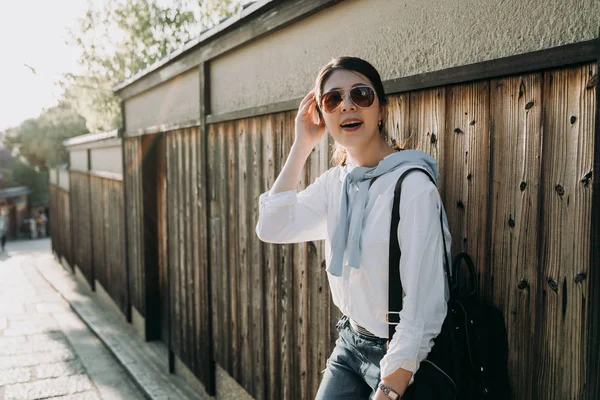 当地的日本女导游依靠木墙等游客完成参观历史名城 年轻女孩戴着太阳镜工作背包放松享受周末的阳光 — 图库照片