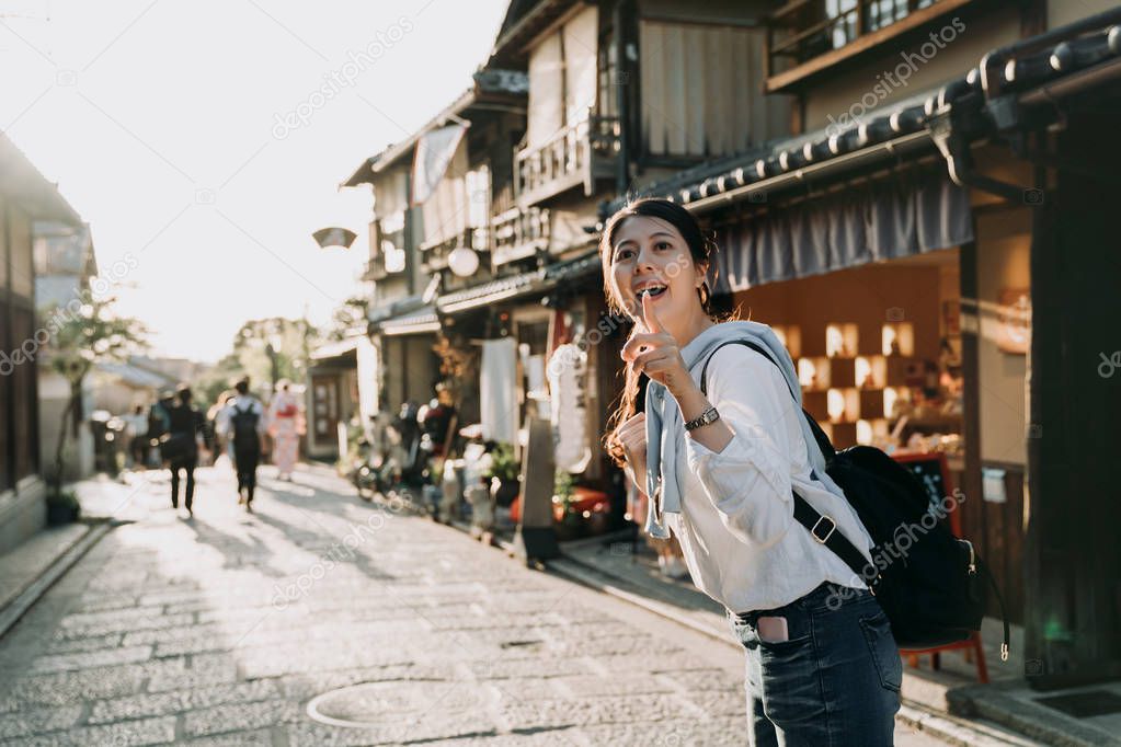 young asian woman backpacker cheerfully smiling pointing to local specialty shop on Hanamikoji Dori. girl tourist stand under sunset in afternoon kyoto japan. happy lady look up to blue sky sunlight.