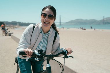 Kız backpacker kiralama seyahat Tur san francisco Kaliforniya ABD Kira. Genç Asyalı kadın yüz kamera neşeyle binicilik bisiklet plajda süre gülümseyerek. Golden gate köprüsü arka planda mavi gökyüzü ile.