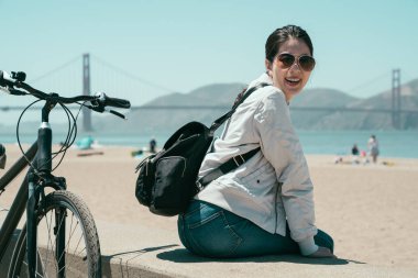 Yerel Asya öğrenci bay güzel yaz görünümü golden gate köprüsü ve okyanus izlerken yanında Bisiklet ile üzerinde oturan bir çanta. Kafa kameraya gülümseyen dönüm güneş gözlüklü genç çekici kız.