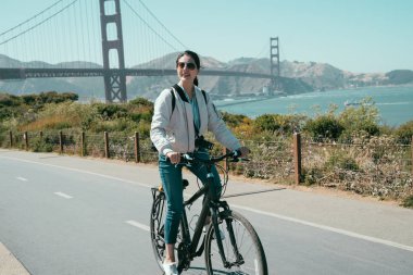rahatlatıcı ve bisiklet turu ABD'de at binme güneşin tadını genç turist kadın güneş gözlüğü. spor kız gezi ziyaret golden state Köprüsü kıyısında Bisiklete binme. Asya Bayan binmek bisiklet yol güneşli