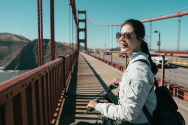 Genç Asyalı kız backpacker yürüyüşüne yaya üzerinde bisiklet ile Golden gate köprüsü ile güneş gözlüğü güneş ışığı altında. Köprüde neşeyle zevk gülümseyen güzel doğa görünümü gezi bisikleti kadın.