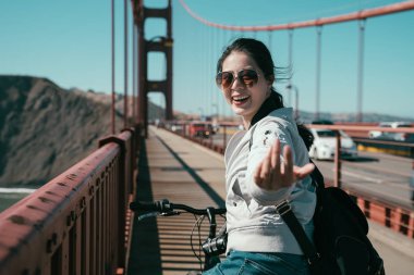 Kız backpacker yaya golden gate köprüsü üzerinde bisiklet sürme. Genç Asyalı çekici Bayan hareketli parmak neşeli gülümseyen söylüyorum onun kayıt seyahat Tur san Francisco ile gelmek için kişileri davet.