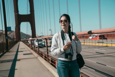 mutlu Genç Asyalı kadın seyahat fotoğrafçı yaya trafik otomobil sürücüsü yolda ile golden gate köprüsü üzerinde yürürken cep telefonu tutuyor. güzel Bayan taşımak kamera arama online harita üzerinde