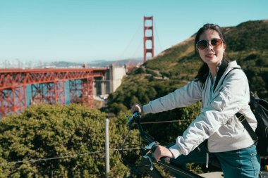 Asya spor kız Bisiklete binme Golden Gate Köprüsü San Francisco yakınındaki açık. güneşli bir gün gezi ünlü Bisiklet Bisiklet sürme güneş gözlüklü genç kadın Simgesel Yapı california. Bayan yüz kamera gülümseyerek..
