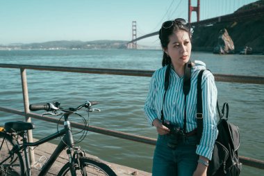 yorgun kadın backpacker kıyı şeridi mavi okyanus deniz kenarında tarafından demir metal Küpeşte üzerinde güvenerek dinleniyor. güneşli bir gün altında arka planda kırmızı golden gate köprüsü. Kız objektif adam eğlence Bisiklet ile rahatlatıcı