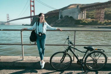 Asyalı kadının güzel manzara görünümü altında güneş ışığı güneşli okyanus denize yakın duran ünlü demir kırmızı golden gate köprüsü üzerinde. Genç kız rahatlatıcı Bisiklet turu kamera taşımak.