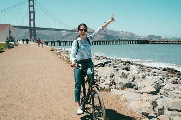 アジアの女性の夏のアメリカ カリフォルニアのゴールデンステート橋で観光を笑顔の自転車に乗っての旅行します 若い女子学生の陽気な日差しの下で青い空に勝利の指サインに手を上げる — ストック写真