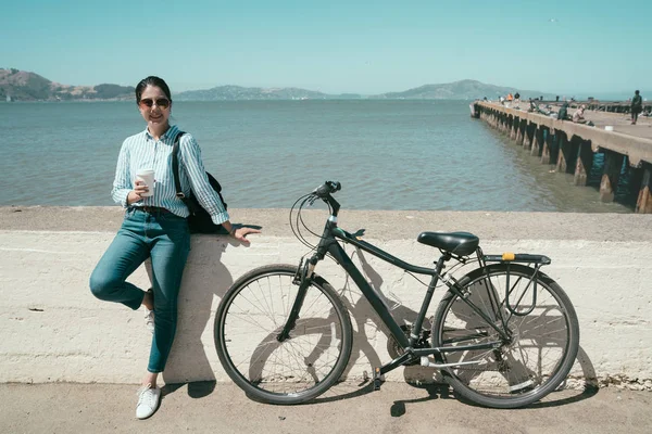 Asya hanım hafta sonu eğlence döngüsü turu açık havada rahatlayın. Genç kız gülümseyen yüz fotoğraf makinesi kahve ile onun tarafından otopark Bisiklet güvenerek dinlenme tutan güneş gözlüğü takıyor. kadın hobi binicilik bisiklet.