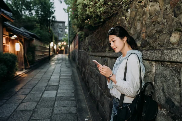 静かな通路の石壁に立っているに沿って待っている地元の日本人女性 チャット オンライン携帯用バックパックの夜の日付の準備ができての笑みを浮かべて携帯電話を保持している女性バックパッカー京都日本します — ストック写真