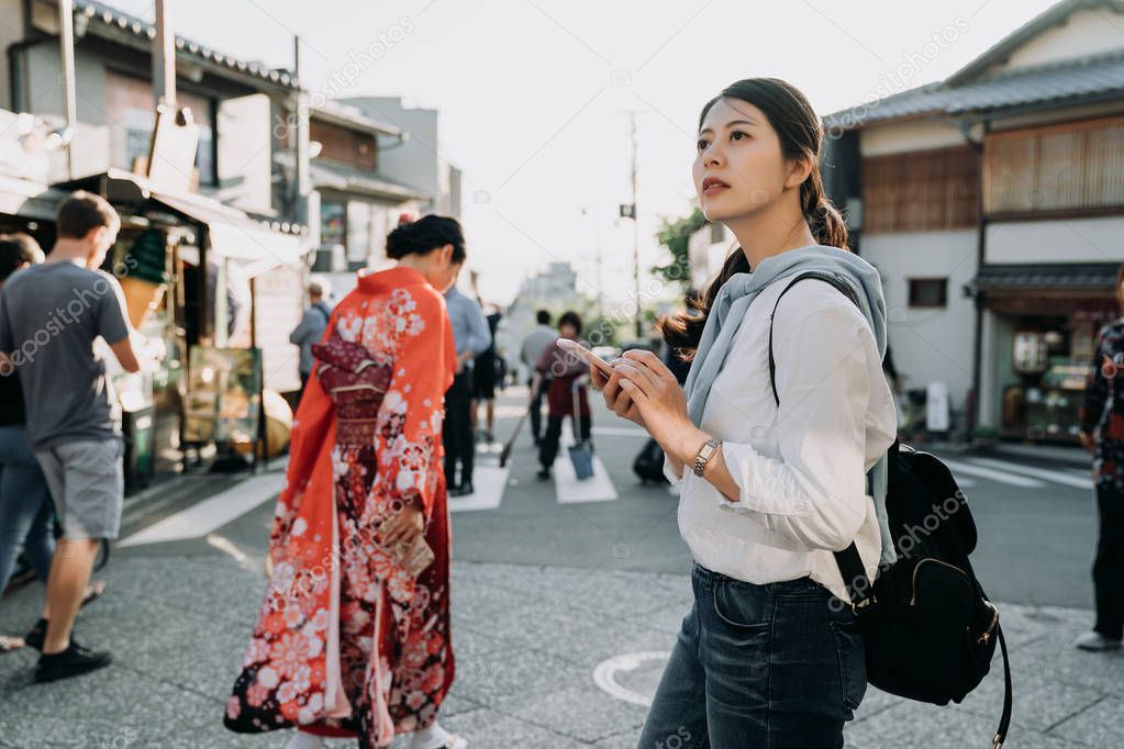 girl traveler hand holding cellphone looking up to road sign finding direction to hotel by online map. beautiful local japanese lady walking in background wearing traditional red floral kimono dress