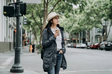 Asyalı kadın açık havada ampul için bekleyen yürürken çay içme yolun karşısına yeşile döner. hasır şapka ve paltosunu kahve şehir palo alto California'da rahatlatıcı tutan genç kız.