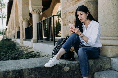 Genç gülümseyen Asyalı kadın öğrenci oturup merdiven açık dış sınıf eski üniversitede binasında. üniversiteli kız Smartphone cep telefonu ile kulaklık ile müzik dinlemek. Eğitim dinlenmek ve rahatlamak kavramı.