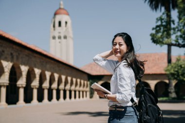 asian college girl standing outdoor going to class at school in summer tour program. elegant smart exchange student study overseas in stanford usa. young woman freshman in new semester cheerful smile clipart