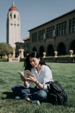 Asya kadın kolej kız okul kitap gülümseyen okuma Üniversitesi'nde bir çanta açık çimenlerin üzerinde oturan güneş keyfini çıkarın. arka planda Bina uzun kule. kadın dili öğrenen öğrenci çalışma yurtdışında.