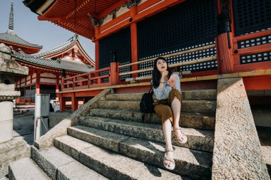 neşeli kız turistik bilgileri akıllı telefon Japon kırmızı ahşap shinto merdivenlerde oturan bir satır kontrol. Genç kadın backpacker kiyomizu dera ziyaret
