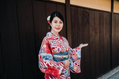 ahşap duvar arka planda tutuyoruz hoş geldiniz el hareketi ile yüz kamera gülümseyen çiçek kimono Japon yerel kız. Bayan söylemek insanlar kyoto Japonya ziyaret gelmek için geleneksel kıyafet konak.