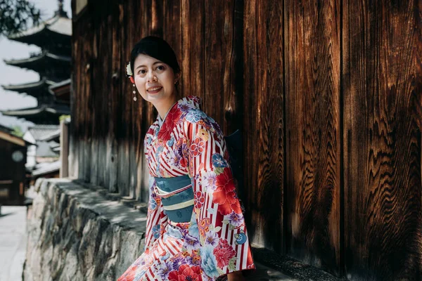 renkli kimono elbise yasaka pagoda içinde geçmiş ile ahşap duvar güvenerek ağaç gölge altında oturan zarif Asyalı kadın. sokak giyim geleneksel bez üzerinde rahatlatıcı gülümseyen genç kız.