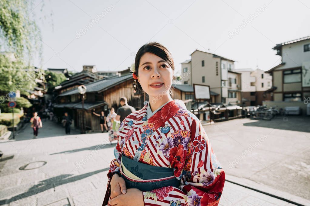 beautiful lady in floral kimono dress look up to blue sky enjoy sunset on street. tourists in background visiting japanese old town kyoto japan. young asian woman in traditional cloth smiling.