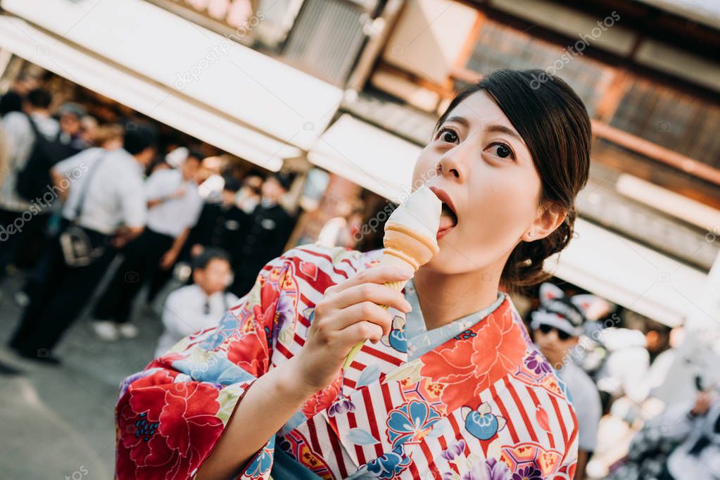 happy japanese local woman in traditional colorful dress eating matcha ice cream standing on teeming street with many tourists in background enjoy sunset while having snack. girl in floral kimono.