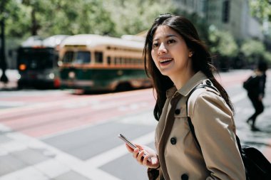 bir kenara Street san Francisco geçiş road yaparken cep telefonu tutan seyir gülümseyen güzel üniversiteli kız. hafif raylı tren arka planda otobüs. mutlu genç kadın eğleniyor akıllı telefon sohbet