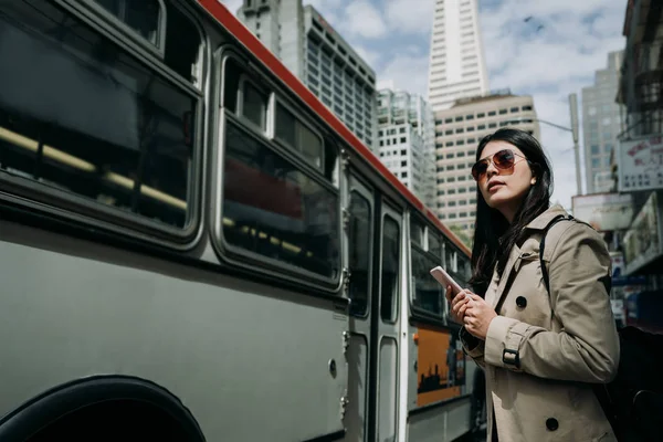 happy woman tourist standing on street near bus stop waiting searching timetable on cellphone. young people technology commute. light rail train close driving pass on road in busy city with blue sky.