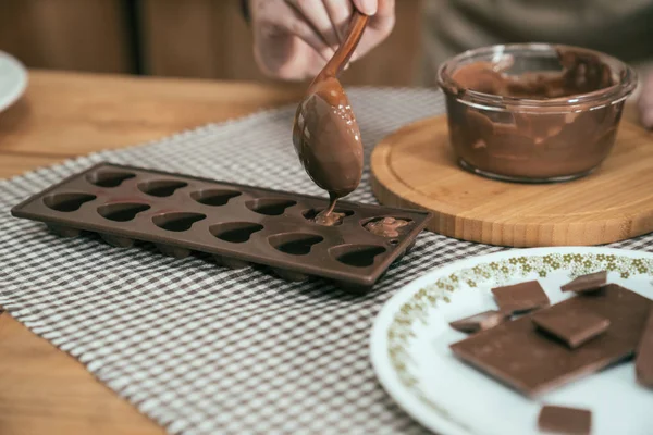 focus cook hands using spoon a silicone dessert mold with melted black chocolate from glass bowl. handmade sweet cocoa fill in heart shape. pieces of chocolate on plate on wooden table in kitchen.