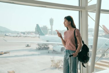 Genç Asyalı kadın backpacker havaalanında uçak için bekliyor. Japon kadın cep telefonu standı pist terminal salonunda Bagaj Taşıma için pencere bakarak tutarak. Seyahat yaşam tarzı kavramı.