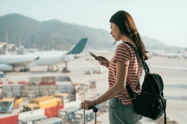güzel seyahat Bayan sırt çantası ve Bagaj bavul için kalkış lounge salonunda yürüyüş ile yan görünüm. cep telefonu sohbet online Uçak piste Window'u yakın duran turist kadın.