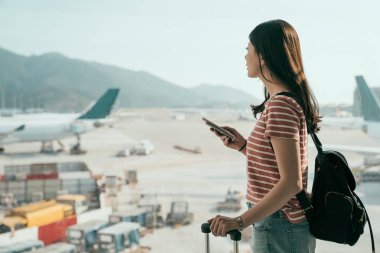 uçuş için bekleyen Havaalanı pencere yakın cep telefonuyla duran genç Asya seyahat kadın yan görünüm. sırt çantası ve Bagaj'lounge alanı pist uçak arayan salonunda güzel kızla