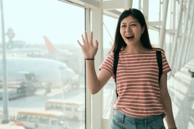 Genç Asyalı kadın, Havaalanı geliş salonunda yürüyüş gümrük kontrolü ver. Yolcu arkasından pistte uçak var. Merhaba arkadaşlar söyleyerek mutlu kız dalgalar el onu almak.