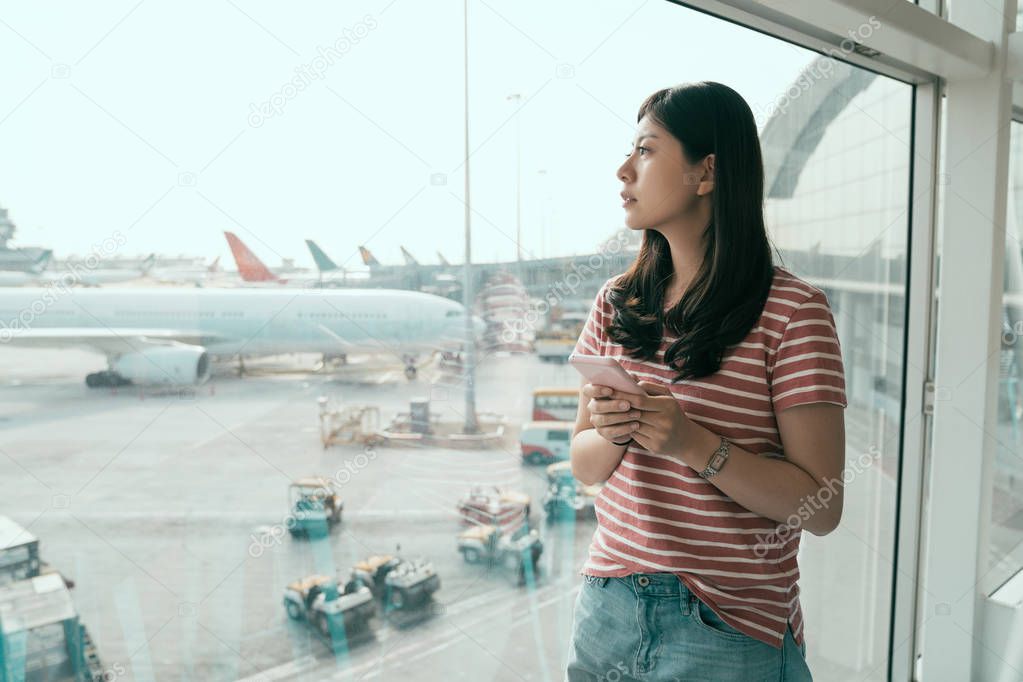 Young chinese woman is standing near window at airport and watching plane before departure. asian girl in casual wear travel summer holiday. female traveler use mobile phone check flight timetable