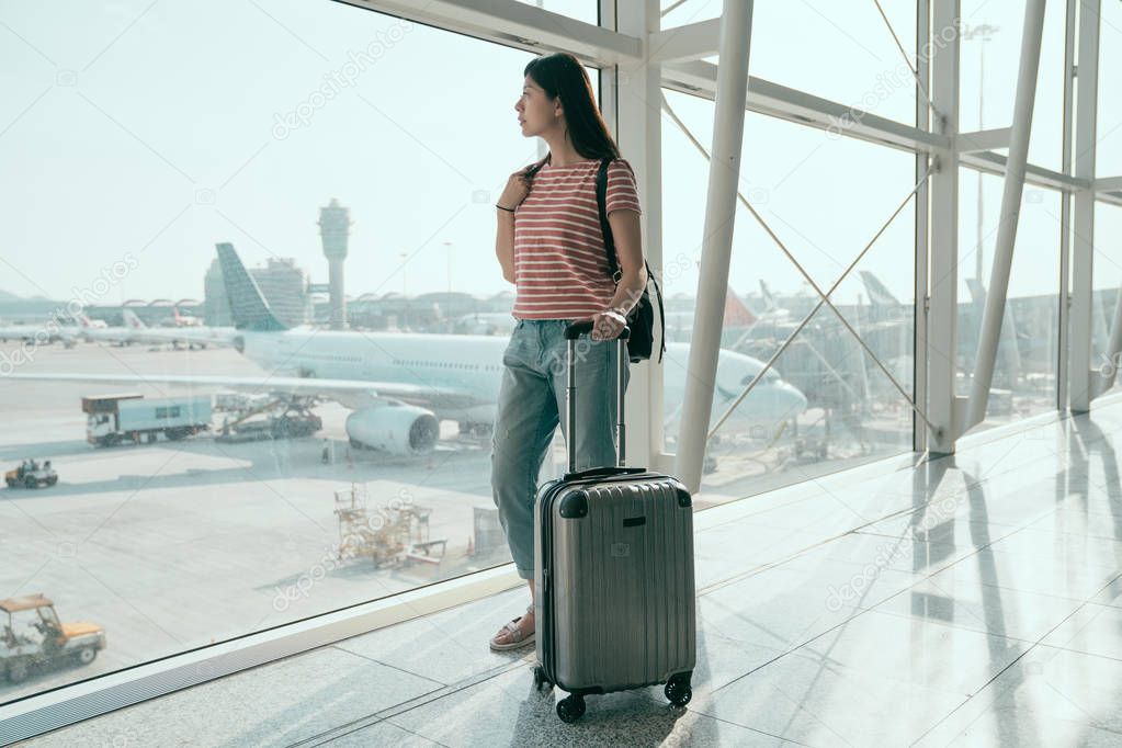 full length of travel female tourist standing with luggage watching outdoor view at airport window. chinese woman at lounge looking at airplanes while waiting at boarding gate before departure.
