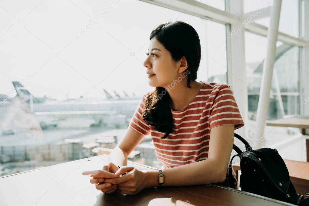 asian woman holding mobile phone in hand waiting for her flight to san francisco at the airport usa. elegant female backpacker looking outside window sitting at table seeing amazing airplane view.