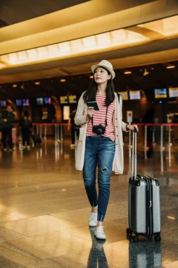 tam uzunlukta genç Asyalı kadın turist çanta ve kalkış kurulu Havaalanı lobi holding pasaport ve biniş kartı onun elinde duran ve seyir için uçuş bilgi Bagaj ile.