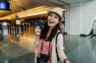 Mutlu genç Asyalı kadın rahat fotoğraf kamera gösteren pasaport ve uçak bileti tutarak Havaalanı lobi terminal kapısı ayakta giymiş. Şapkalı Kız yurt dışına gidiş yaz tatile seyahat.