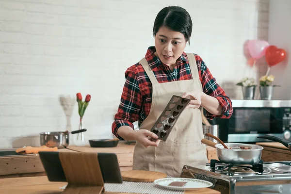 young woman cook in apron recording video tutor teaching how to handmade chocolate on valentine day. girl showing heart mold to digital tablet on table in modern wooden kitchen while melting cocoa.