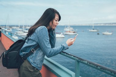 öğleden sonra plaj Koyu'ndaki tarihi bekleyen cep telefonu tutan genç yerel Asya Bayan. kız sırt çantası ile iletişim kurma arkadaş sohbet cep telefonu okyanus tekne görmek eski Fisherman's Wharf görmek.
