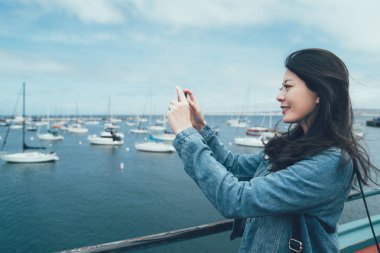 yan görünüm genç Asyalı kadın turist alarak fotoğraf yat ile akıllı telefon önünde eski Fisherman's Wharf monterey koltukta. tekneler çevre beach bay üzerinde cep telefonu ayakta tutan gülümseyen kadın Gezgin
