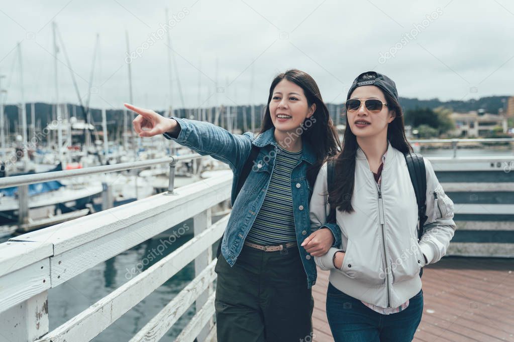 two young asian female friends walking outdoors in harbor port in Old Fisherman's Wharf monterey. happy sisters holding hands enjoying life relaxing sightseeing outside. girl finger showing view