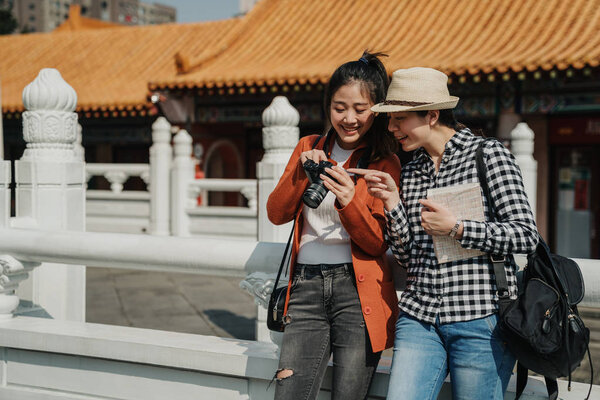 Two happy female tourists checking photos on camera leaning on chinese traditional building stone railings. young girl point finger showing friend laughing smiling chatting hold bag and guide book