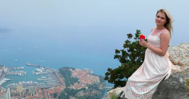 岩の上に座って 彼女の髪に吹く風のゴージャスなピンクのドレスで美しい若いブロンドの女性の肖像画 ビデオのモナコの空撮 — ストック動画